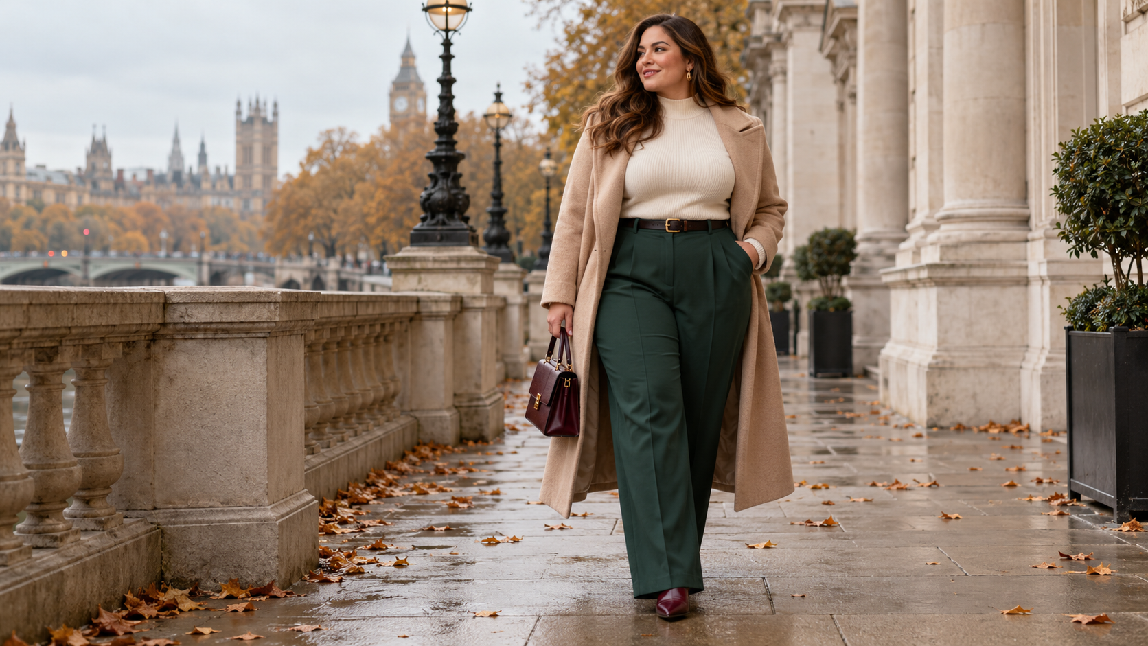 Plus-size woman wearing tailored green trousers, a cream knit and camel coat on an autumn London riverside walk
